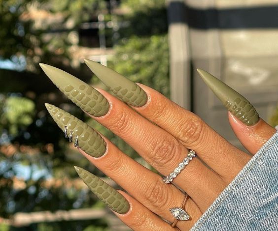 Long stiletto nails with olive green crocodile textures and nail piercings. 
